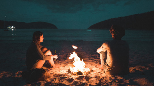 Two people sitting by a camp fire on the beach at night